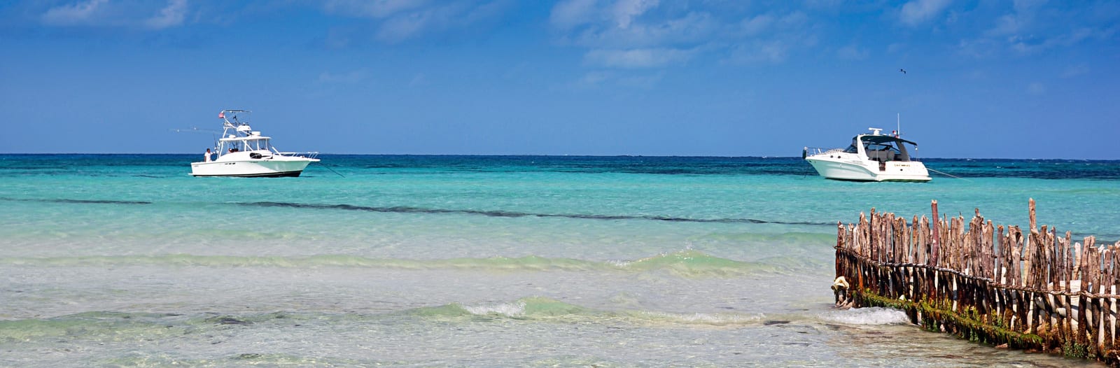 Turquoise waters in Cancun with two boats floating near a rustic wooden fence in the ocean, under a clear blue sky, capturing the tranquil Caribbean seascape at this popular vacation spot. | Houfy — No Fees, Book Direct