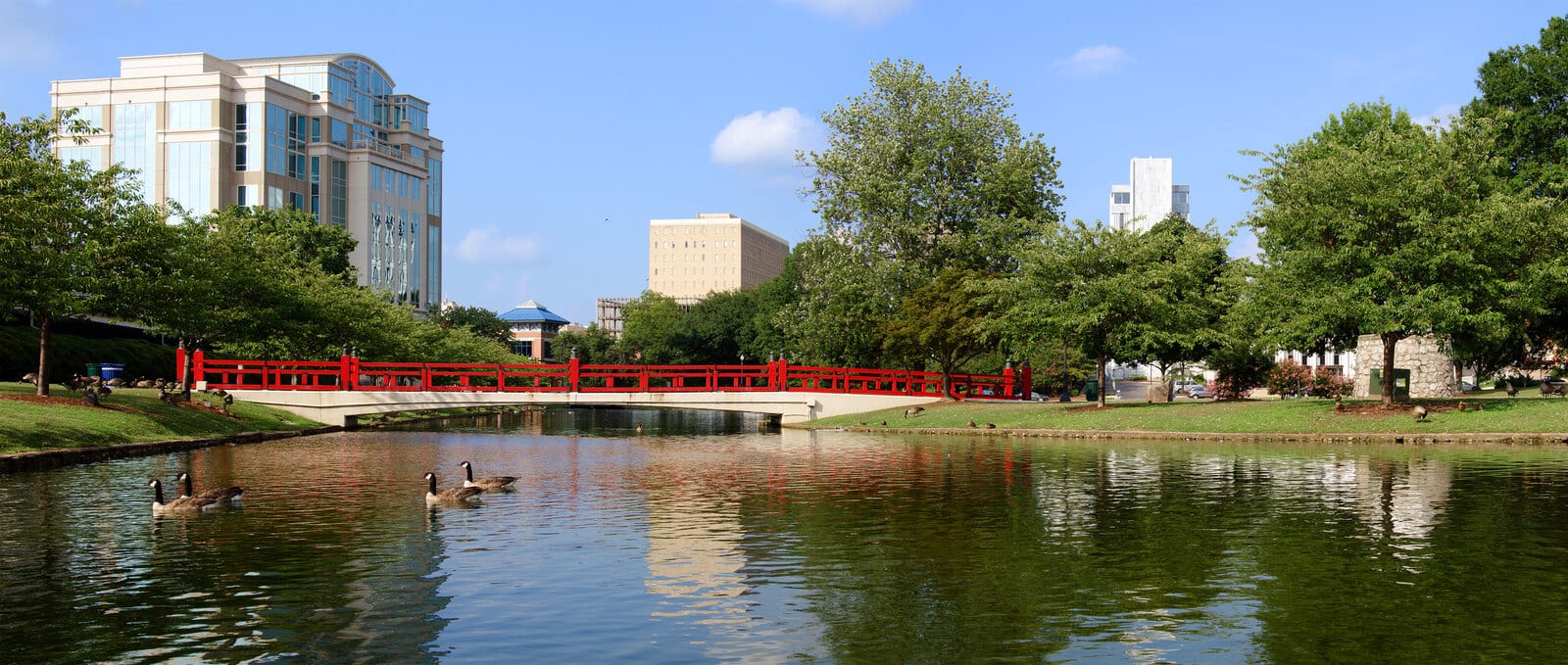 Red bridge over a serene pond with geese, surrounded by lush trees and modern architecture in Huntsville, Alabama, offering a peaceful blend of nature and urban scenery. | Houfy — No Fees, Book Direct