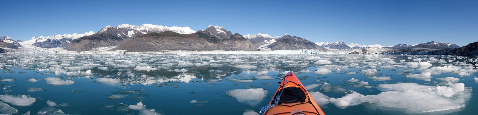 Kayaking through icy waters with a view of majestic snow-capped mountains in Glacier Bay National Park, Alaska; fragments of ice float serenely, reflecting a clear blue sky. | Houfy — No Fees, Book Direct