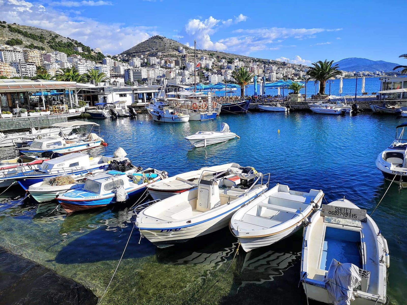 Panoramic view of Sarandë harbor on the Albanian Riviera, with crystal-clear turquoise water, moored boats, and white Mediterranean buildings terracing up the hillside — book a fee-free Albania vacation rental on Houfy | Houfy — No Fees, Book Direct