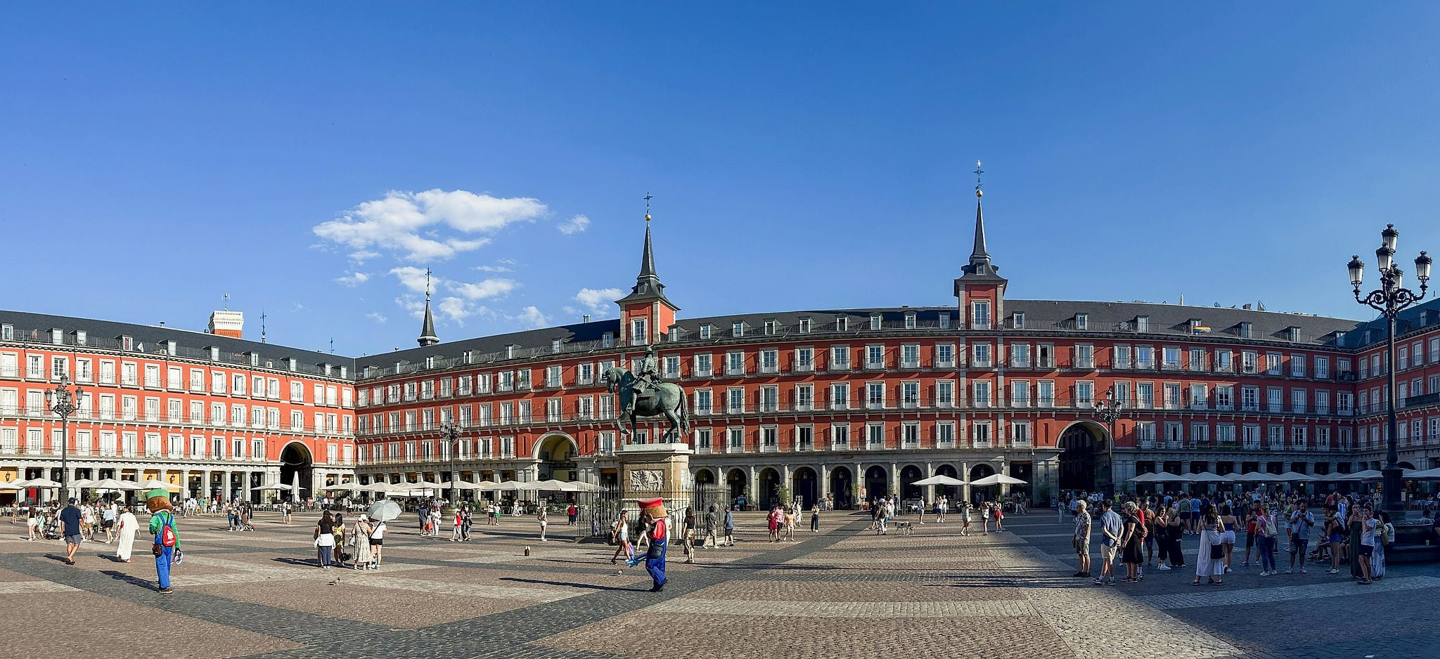 Panoramic view of Plaza Mayor in Madrid, Spain, with vibrant terracotta-red Hapsburg facades, wrought-iron balconies, stone arcades, the bronze equestrian statue of King Philip III, and tourists on cobblestones under a clear blue sky — book a fee-free Madrid vacation rental on Houfy | Houfy — No Fees, Book Direct