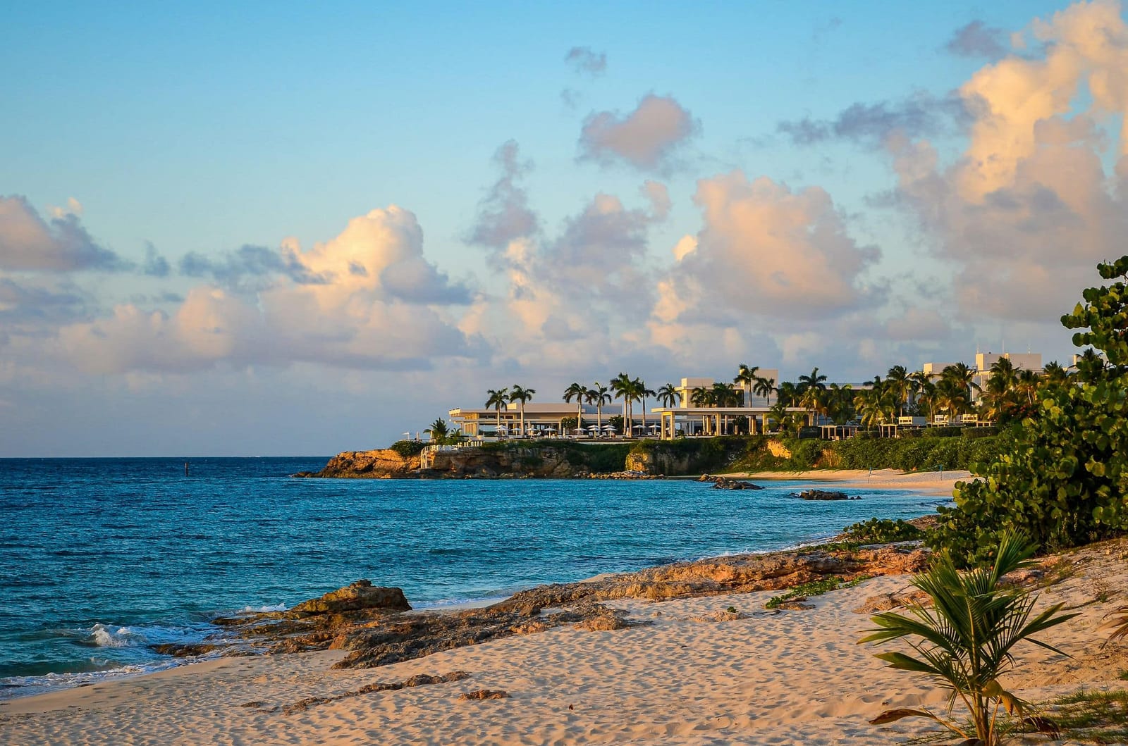 Luxury beachfront villas perched on a rocky cliff in Anguilla at golden hour, with a pristine white sand beach and turquoise Caribbean Sea — book a fee-free Anguilla vacation rental on Houfy | Houfy — No Fees, Book Direct