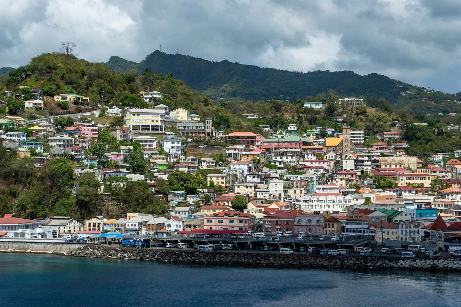 Colorful Caribbean coastal town with terracotta rooftops and pastel buildings cascading down a lush tropical hillside over a calm blue harbor — book a fee-free Antigua and Barbuda vacation rental on Houfy | Houfy — No Fees, Book Direct