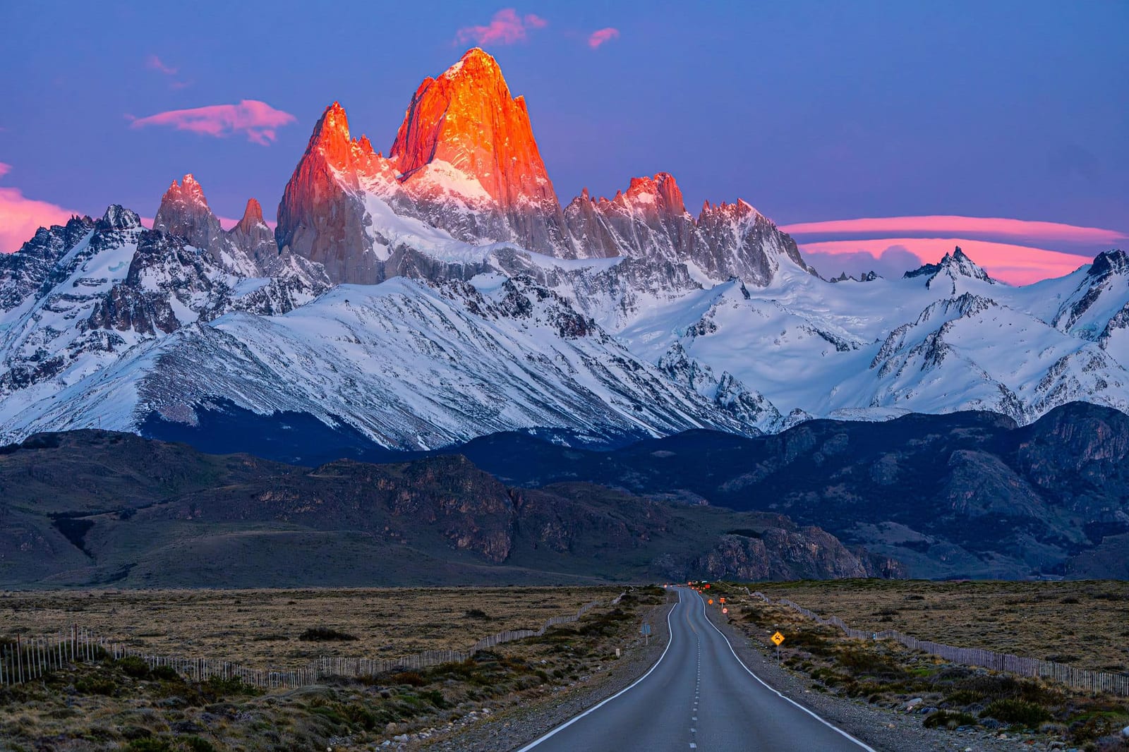 Dramatic sunrise over Monte Fitz Roy in Argentine Patagonia, with a long straight road leading toward snow-capped granite peaks glowing orange at golden hour — book a fee-free Argentina vacation rental on Houfy | Houfy — No Fees, Book Direct