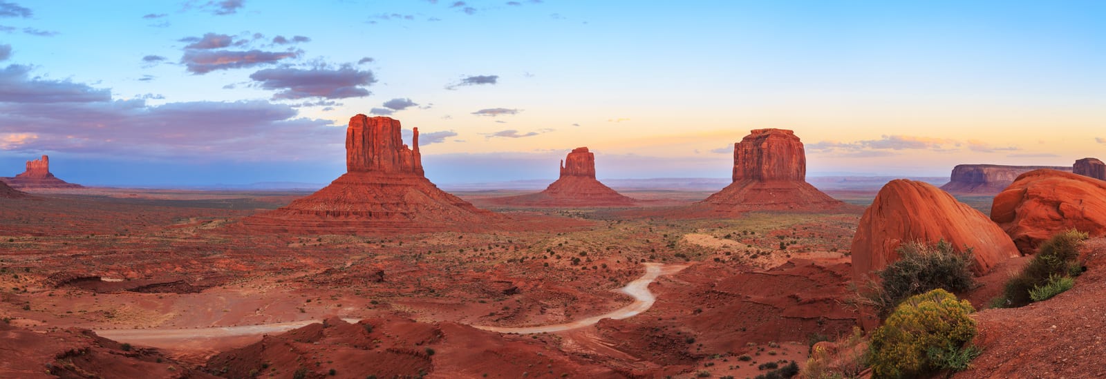 Monument Valley's iconic buttes rise against a pastel sunset sky, surrounded by red desert terrain. The winding dirt road offers a distinct perspective of this timeless American landscape. | Houfy — No Fees, Book Direct