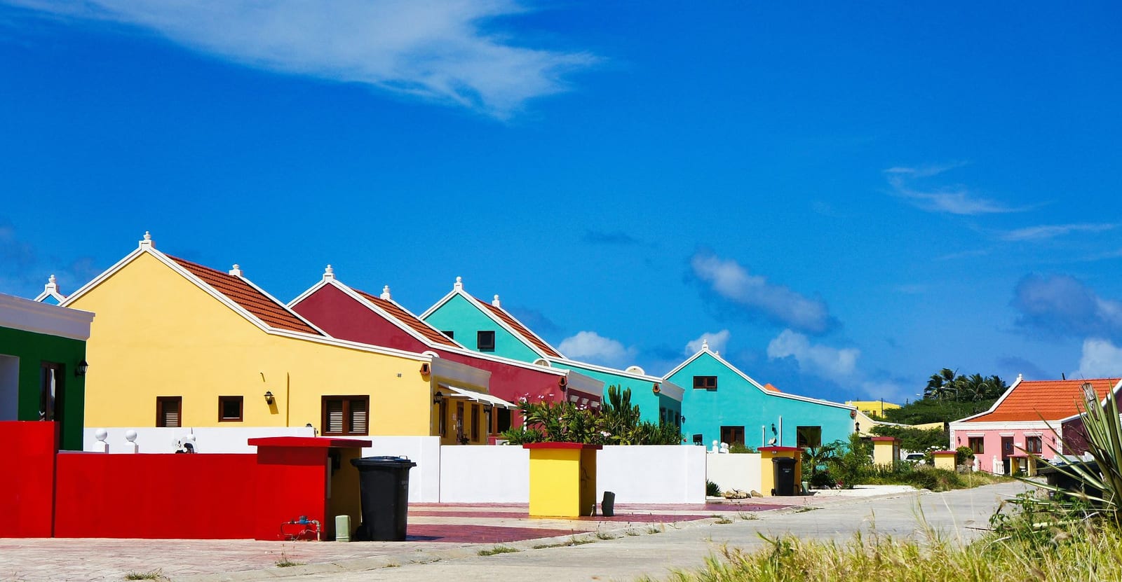 Row of vibrant yellow, turquoise, and maroon Dutch Caribbean vacation villas with white-trimmed gabled roofs under a brilliant blue Aruba sky — book a fee-free Aruba vacation rental on Houfy | Houfy — No Fees, Book Direct