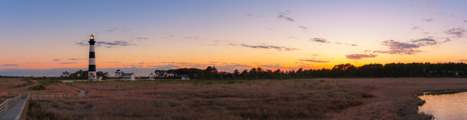 Cape Hatteras Lighthouse stands tall at sunrise, surrounded by marshland and a serene orange sky, offering a tranquil scene unique to the Outer Banks of North Carolina. | Houfy — No Fees, Book Direct