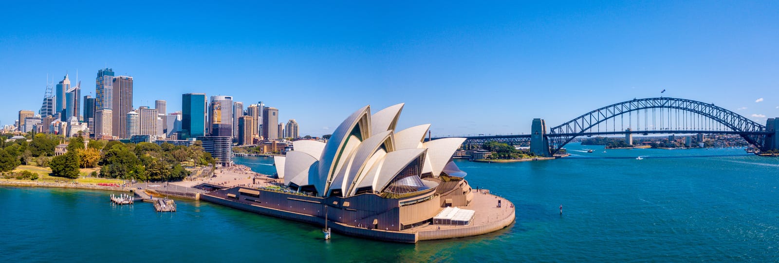 Panoramic view of Sydney Harbour with the iconic Sydney Opera House and Harbour Bridge under a brilliant blue sky — book a fee-free Australia vacation rental on Houfy | Houfy — No Fees, Book Direct