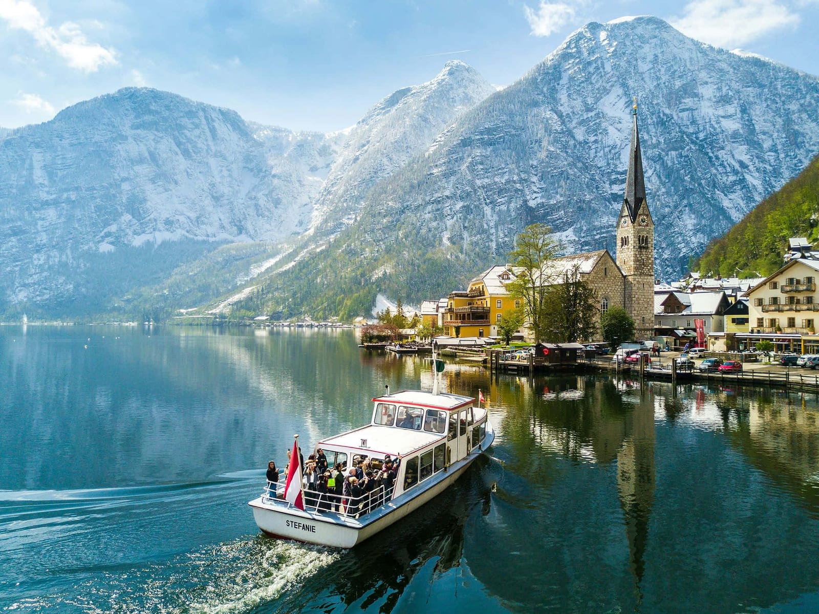 Iconic view of Hallstatt village on Lake Hallstatt, Austria, with a passenger boat, Alpine church spire, traditional yellow facades, and snow-capped Dachstein mountains — book a fee-free Austria vacation rental on Houfy | Houfy — No Fees, Book Direct