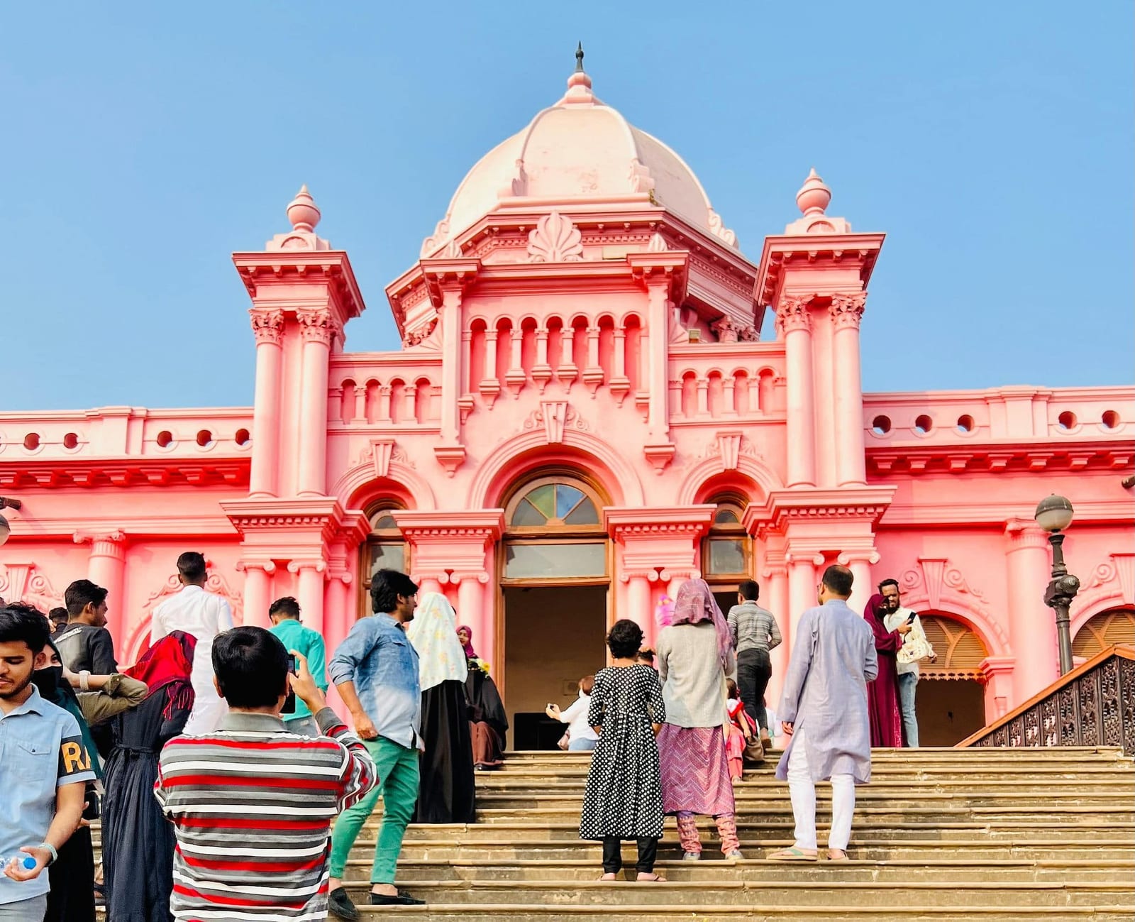 Tourists on the grand staircase of Ahsan Manzil, the iconic Pink Palace in Old Dhaka, Bangladesh, featuring its vibrant bubblegum-pink Indo-Saracenic dome and arched facade under a clear blue sky — book a fee-free Bangladesh vacation rental on Houfy | Houfy — No Fees, Book Direct