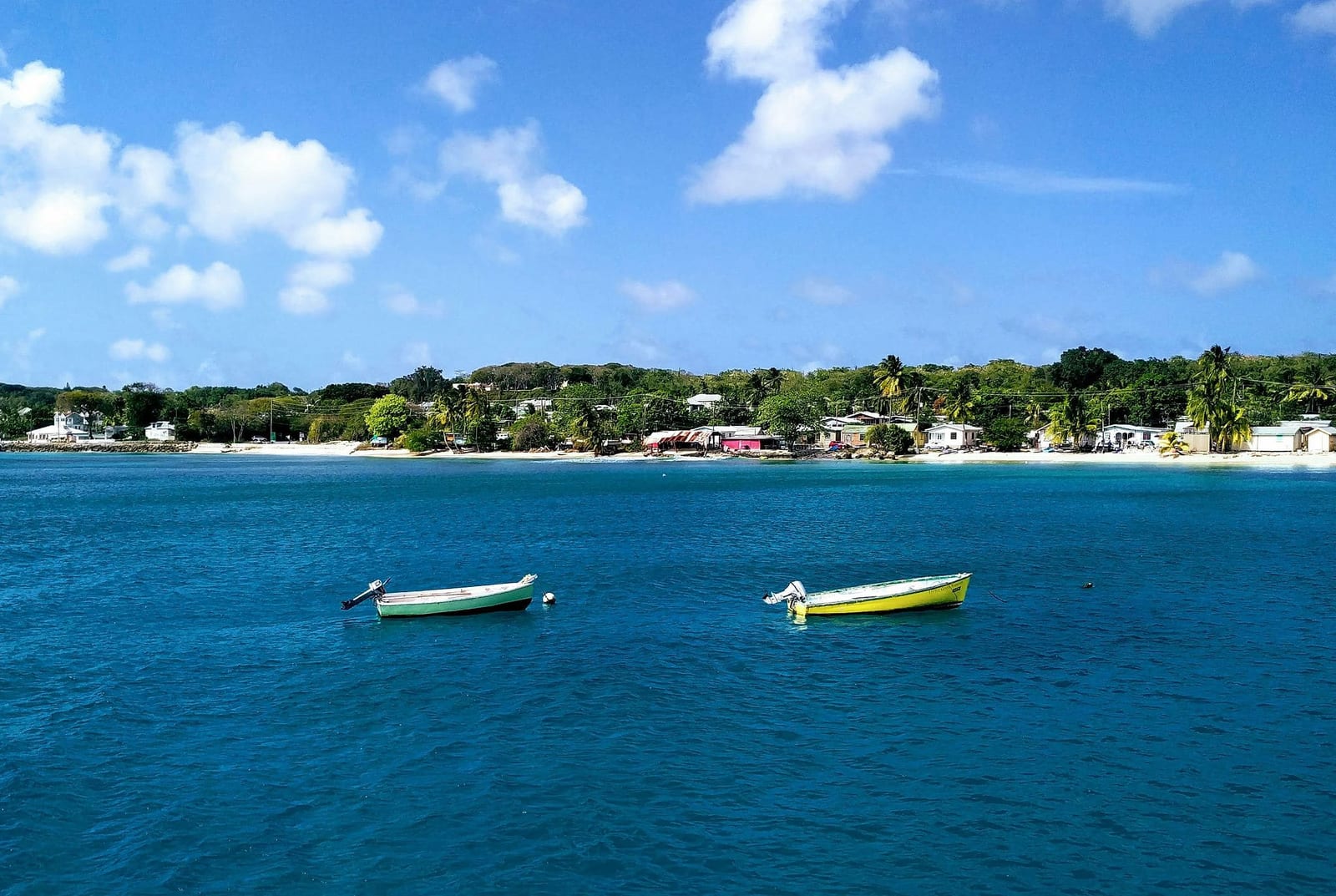 Two colorful traditional fishing boats anchored in crystal-clear turquoise Caribbean waters off a lush Barbados coastline, with white sand beach, swaying palm trees, and a quiet coastal village — book a fee-free Barbados vacation rental on Houfy | Houfy — No Fees, Book Direct