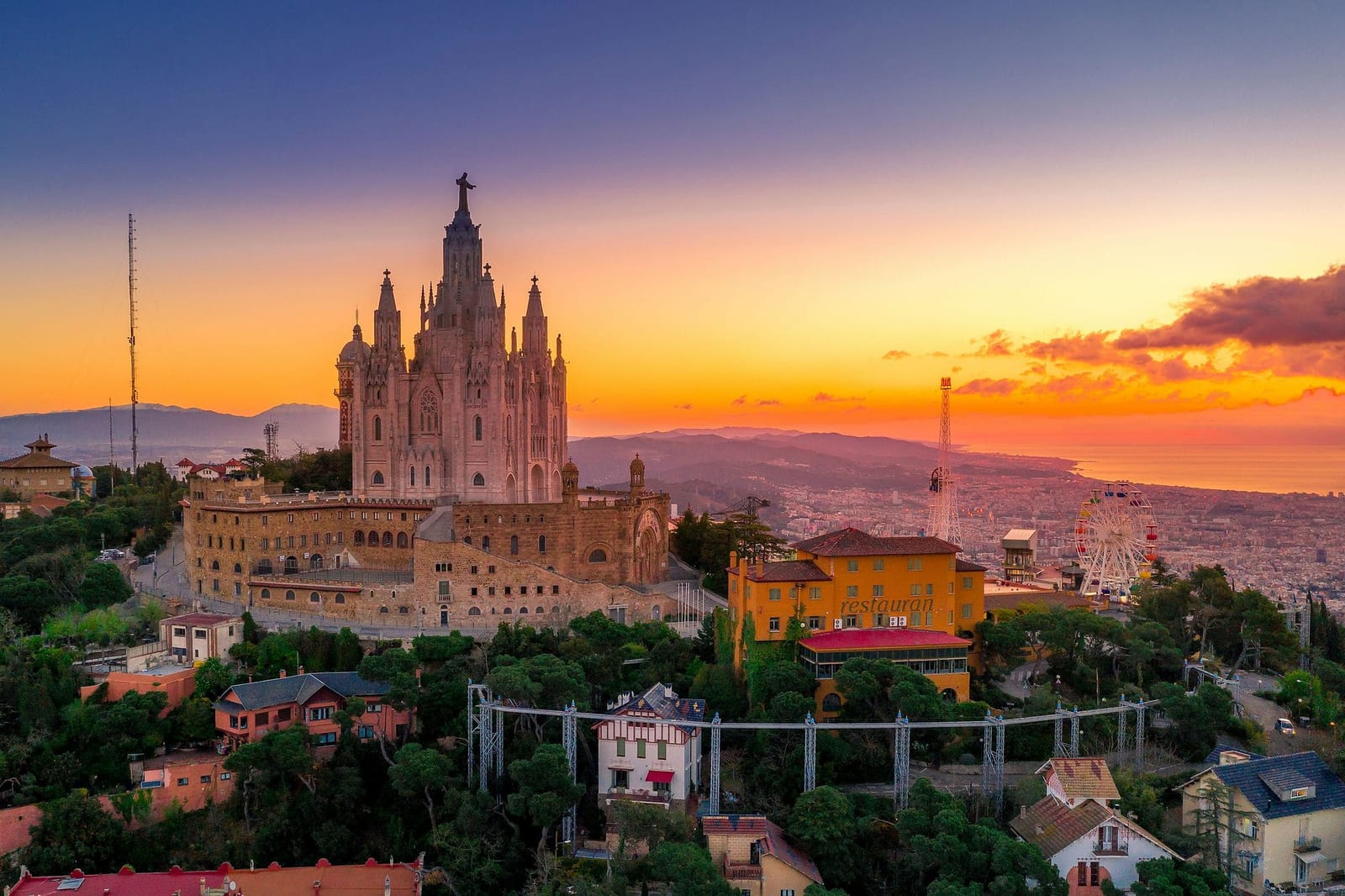 Panoramic golden hour view from Mount Tibidabo in Barcelona, Spain, featuring the Neo-Gothic Sagrat Cor cathedral with bronze Jesus statue, the Tibidabo amusement park, and a fiery violet and orange sunset sky over the Mediterranean — book a fee-free Spain vacation rental on Houfy | Houfy — No Fees, Book Direct