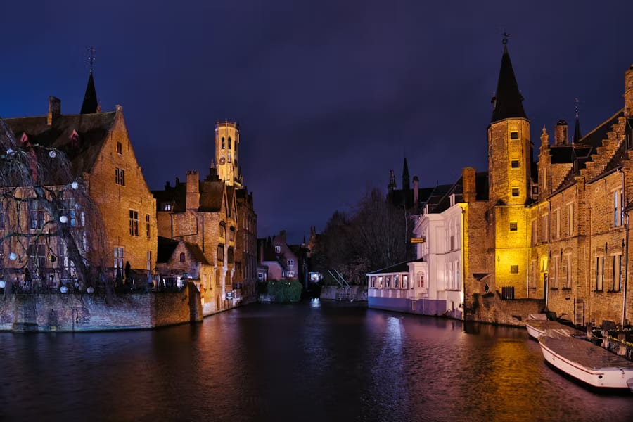 Enchanting blue hour view of Rozenhoedkaai canal in Bruges, Belgium, with illuminated Flemish Gothic brick buildings, the medieval Belfry tower, and golden reflections shimmering on the water — book a fee-free Belgium vacation rental on Houfy | Houfy — Book Direct