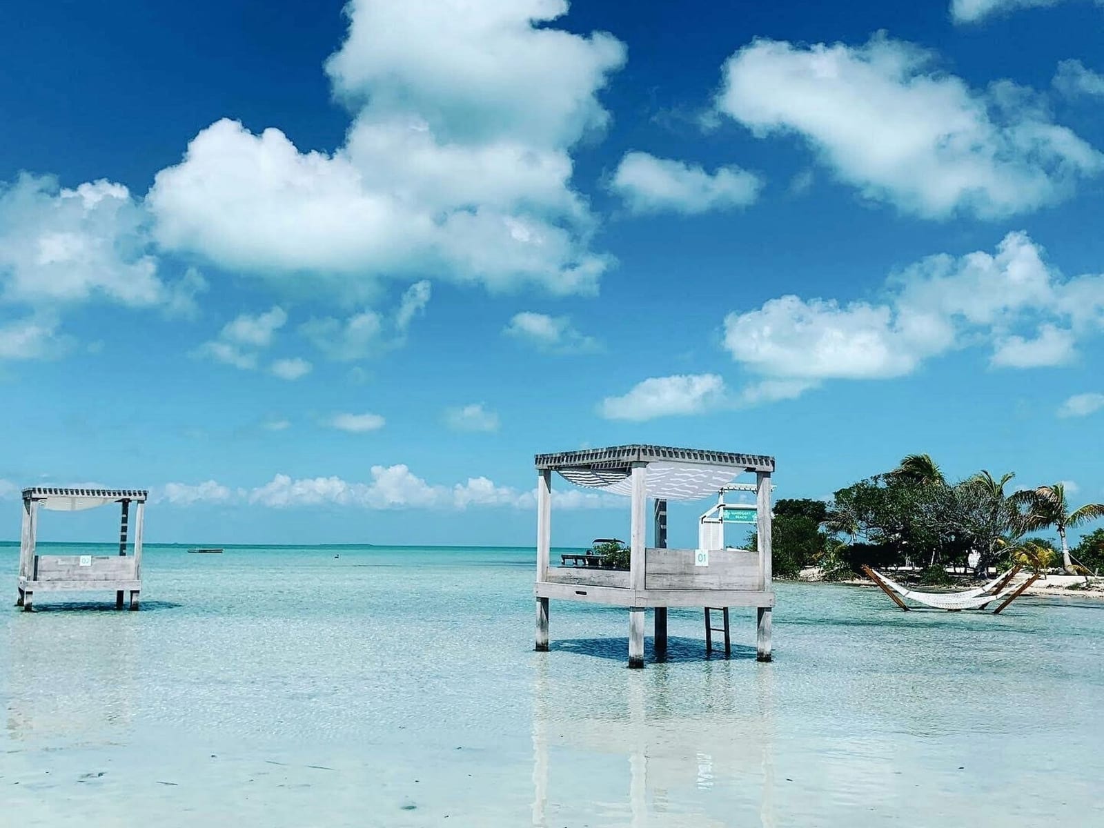 Weathered wooden overwater cabanas on stilts in the crystal-clear shallow turquoise waters of Secret Beach, Ambergris Caye, Belize, with white hammocks and palm-lined shore in the background — book a fee-free Belize vacation rental on Houfy | Houfy — No Fees, Book Direct