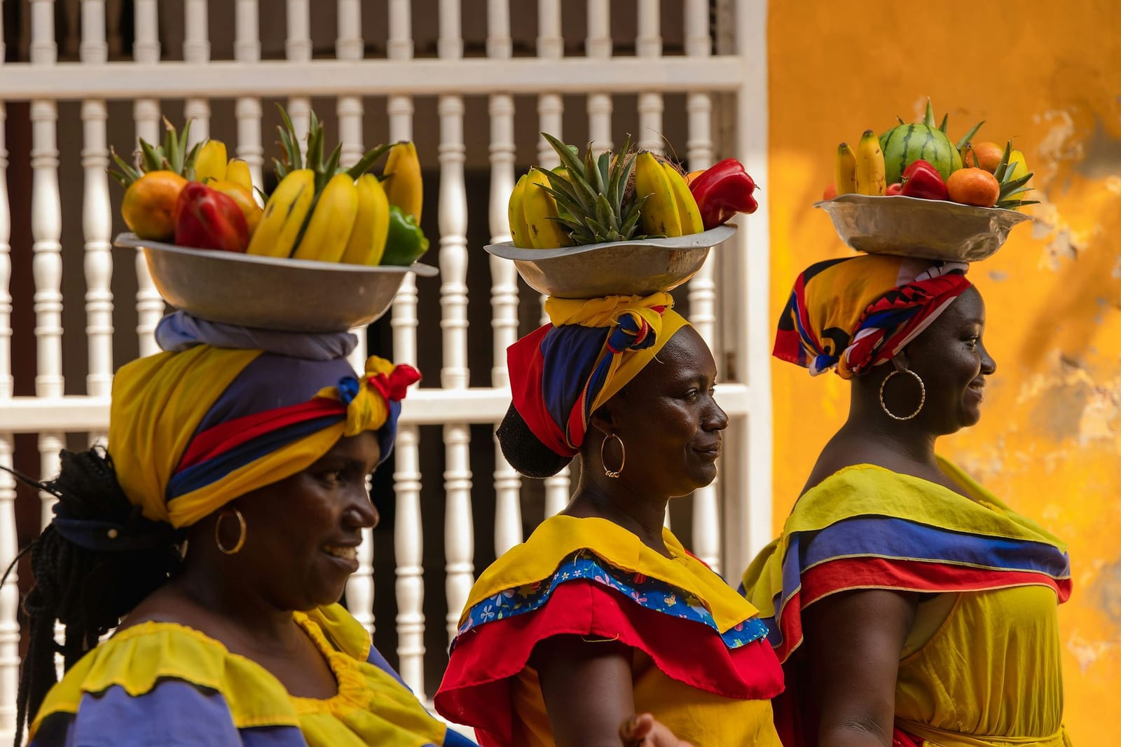 Three women in vibrant traditional yellow, blue, and red ruffled dresses balancing overflowing bowls of tropical fruit on their heads against a sun-drenched colonial wall — book a fee-free Benin vacation rental on Houfy | Houfy — No Fees, Book Direct