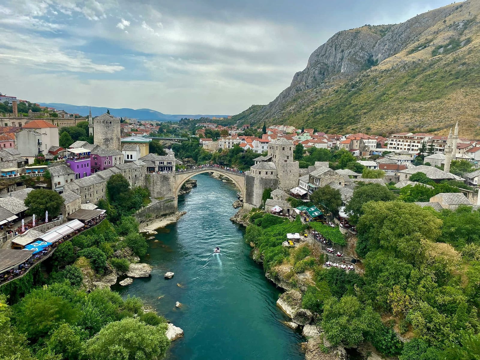 High-angle view of Mostar's UNESCO-listed Stari Most (Old Bridge) arching over the brilliant emerald-green Neretva River, flanked by Ottoman stone towers, terracotta-roofed old town buildings, mosque minarets, and cliffside cafe terraces against a backdrop of limestone mountains — book a fee-free Bosnia and Herzegovina vacation rental on Houfy | Houfy — No Fees, Book Direct