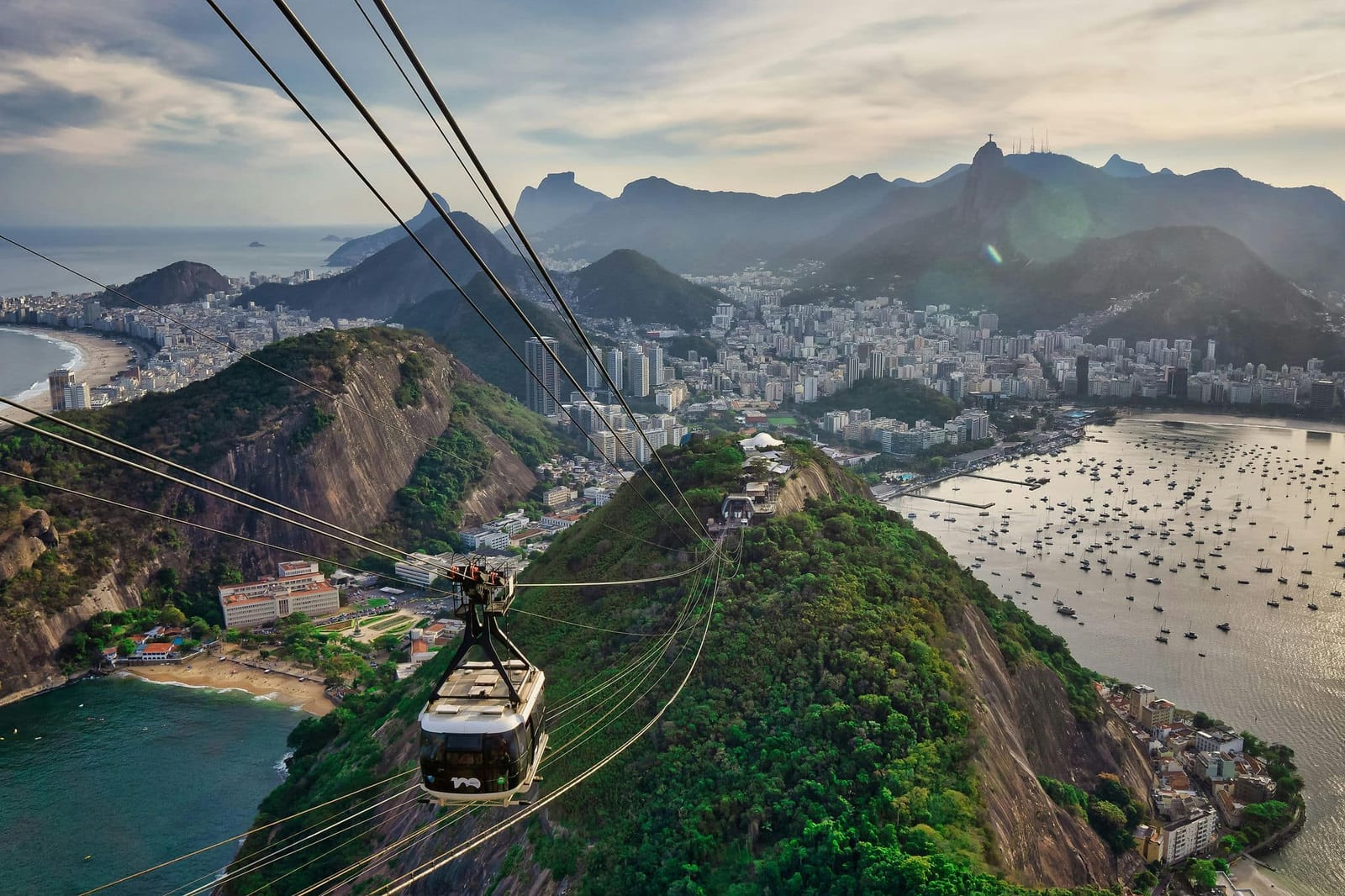 Aerial golden hour view of Rio de Janeiro from the Sugarloaf Mountain cable car, with Botafogo Bay dotted with sailboats, the secluded curve of Praia Vermelha, Copacabana Beach in the distance, and Christ the Redeemer silhouetted on Corcovado — book a fee-free Brazil vacation rental on Houfy | Houfy — No Fees, Book Direct