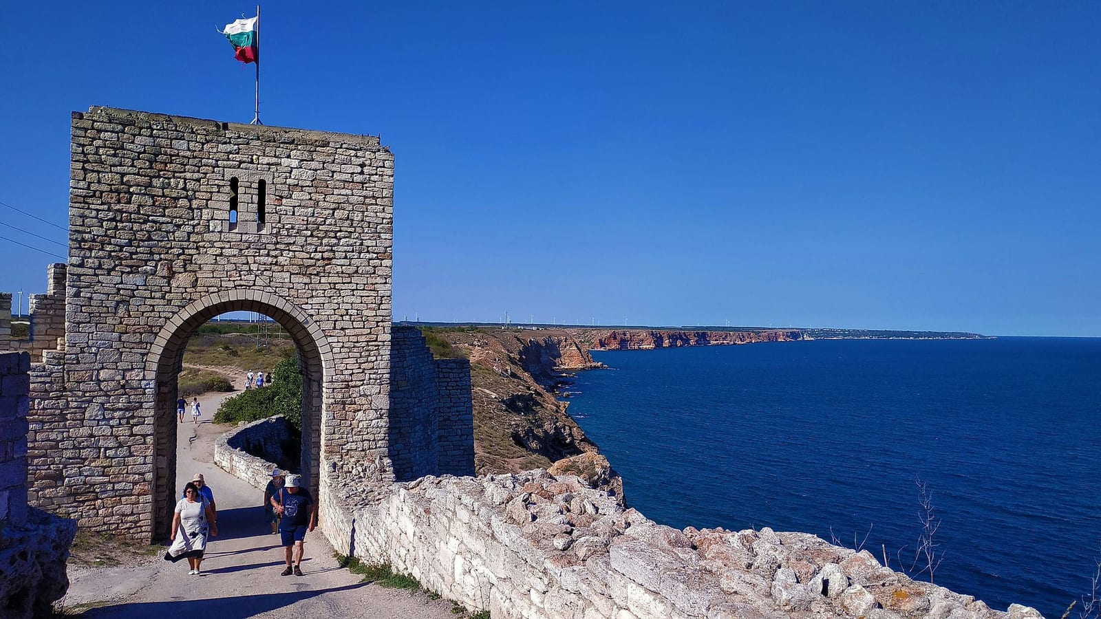 Tourists walking through a medieval limestone archway at Cape Kaliakra, Bulgaria, with a Bulgarian flag flying atop the tower, dramatic reddish-brown cliffs plunging into the deep cobalt Black Sea, and a cloudless cerulean sky — book a fee-free Bulgaria vacation rental on Houfy | Houfy — No Fees, Book Direct