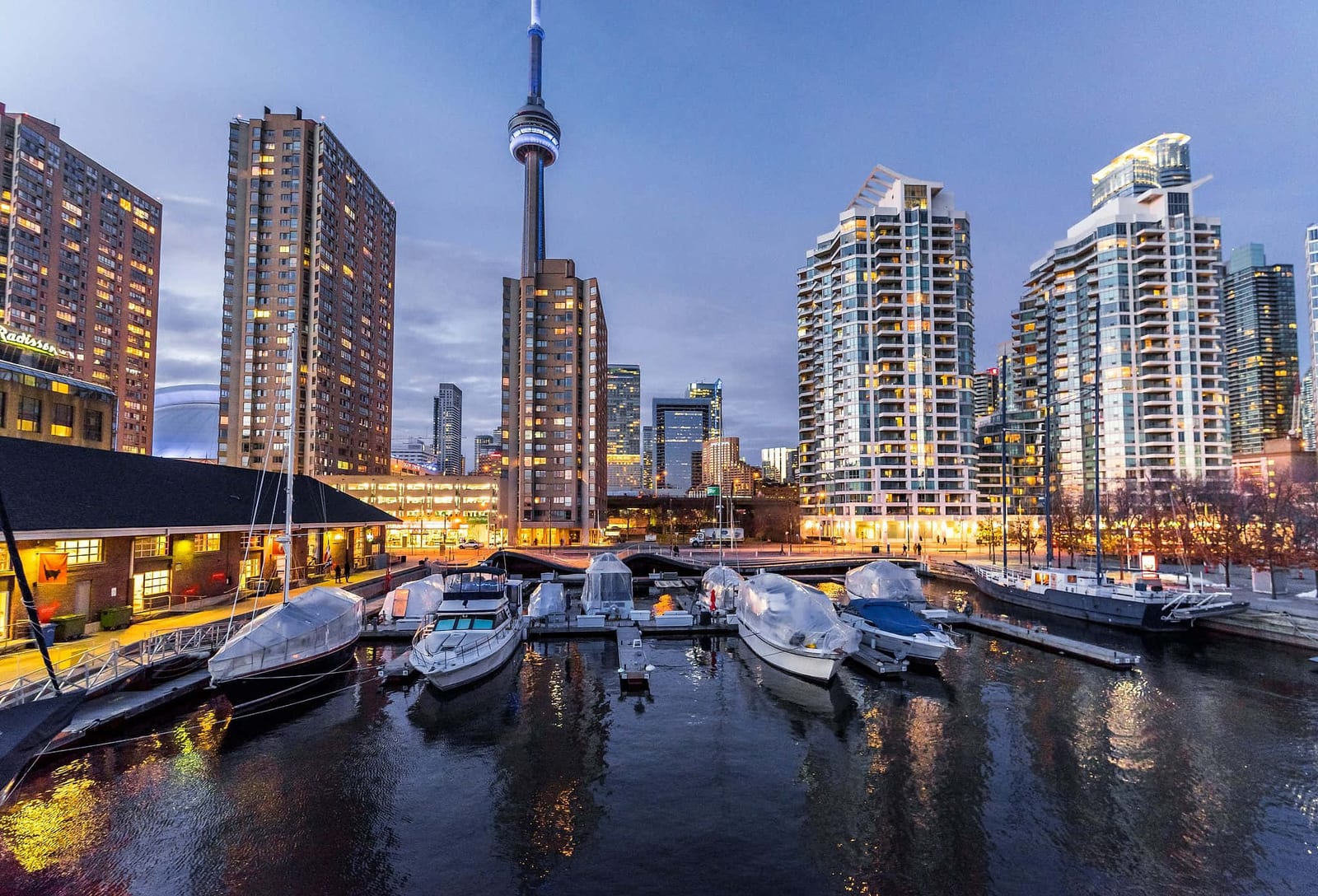 Blue hour view of Toronto's Harbourfront marina, Canada, with the iconic CN Tower illuminated against a deep indigo sky, modern glass condo towers glowing gold, and boats reflected in the calm dark harbor water — book a fee-free Canada vacation rental on Houfy | Houfy — No Fees, Book Direct