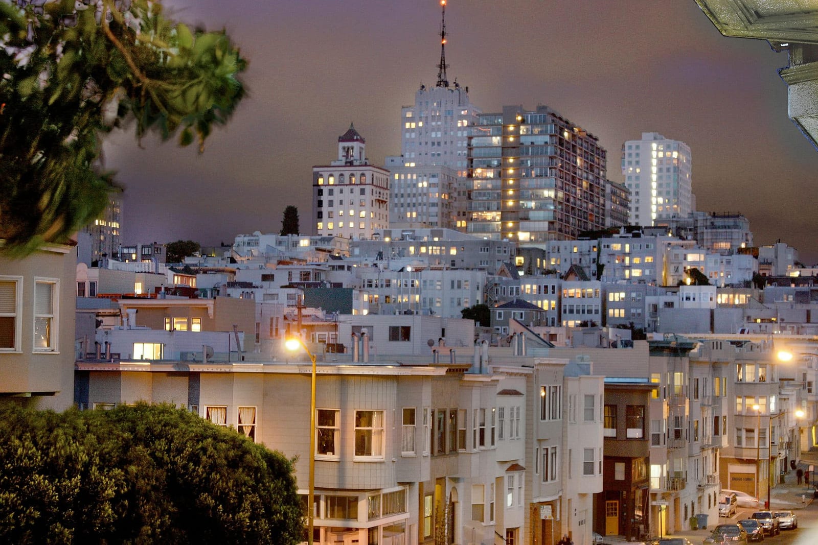 Twilight view of the San Francisco skyline from a fee-free city vacation rental on Houfy, featuring warm glowing city lights, historic bay-window row houses, and downtown skyscrapers at dusk. | Houfy — No Fees, Book Direct