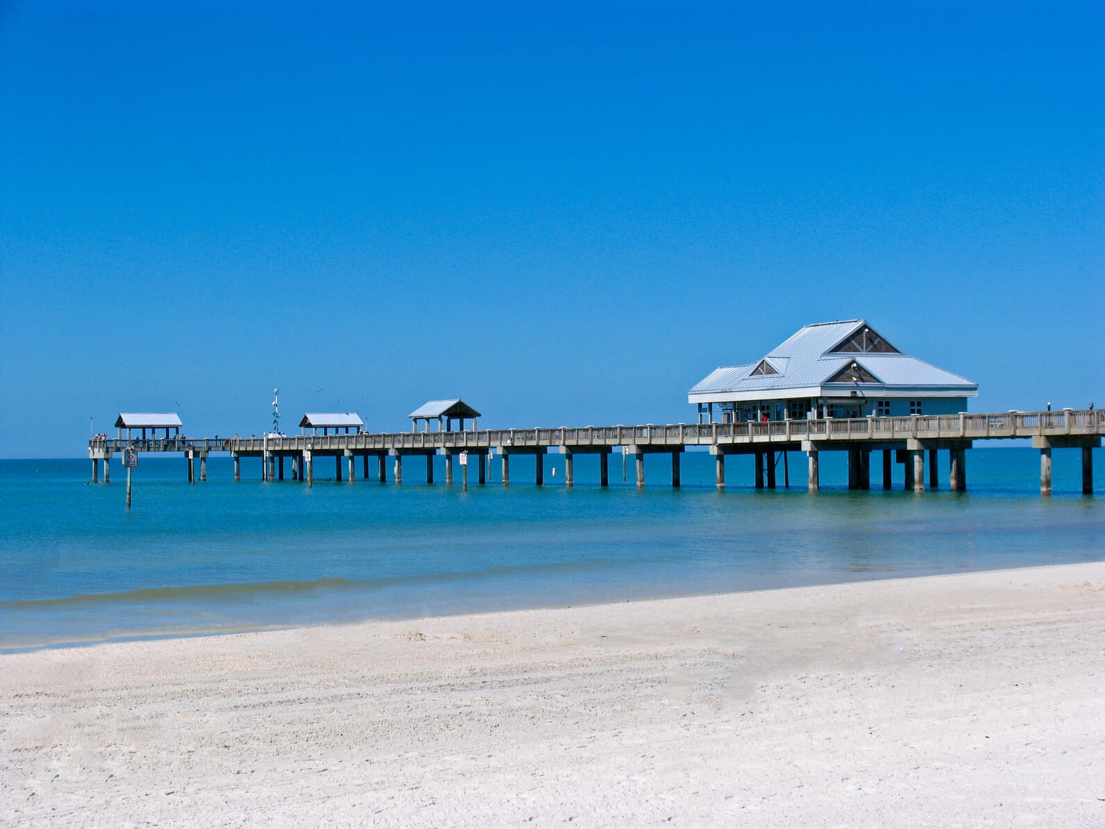 Fort Myers Beach pier stretches into the calm, blue Gulf of Mexico, showcasing its distinct pavilion-style architecture under a cloudless sky, highlighting the pristine sandy shoreline. | Houfy — No Fees, Book Direct