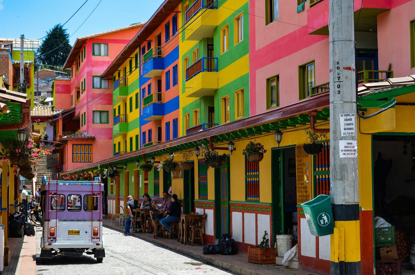 Vivid street scene in Guatapé, Antioquia, Colombia, with multi-story buildings in hot pink, sunshine yellow, lime green, and cobalt blue, ornate zócalo bas-relief facades, flower-basket balconies, terracotta tile rooftops, locals at wooden cafe tables, and a purple moto-taxi on cobblestone streets under a brilliant midday sky — book a fee-free Colombia vacation rental on Houfy | Houfy — No Fees, Book Direct