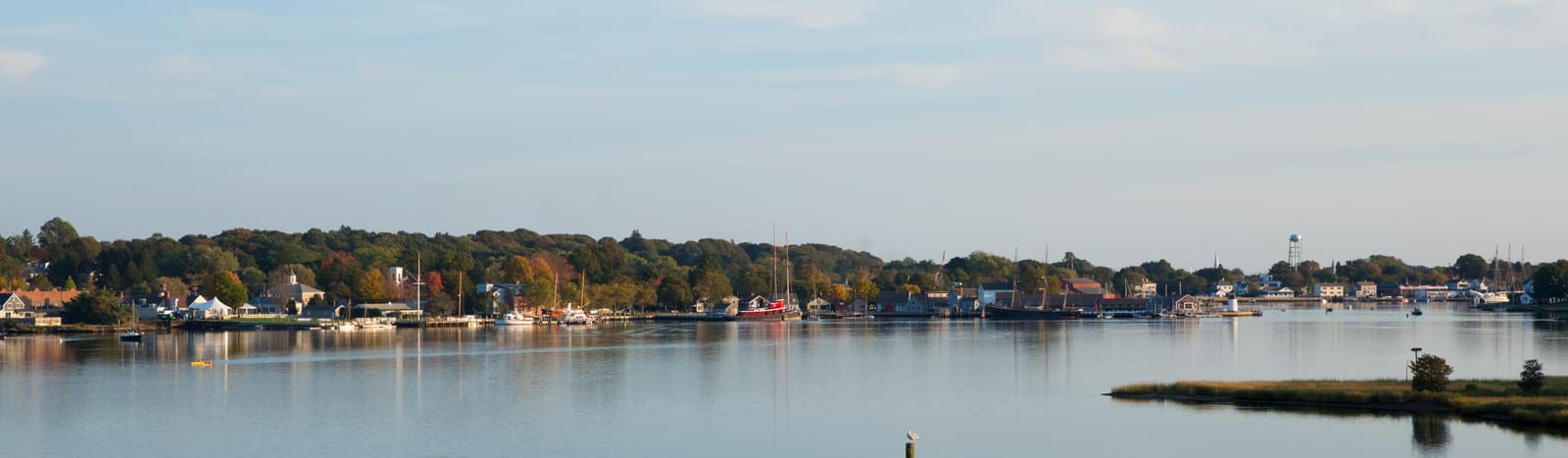 Idyllic waterfront scene in Christiansø, Denmark, featuring quaint houses, a calm harbor with boats, and a lighthouse visible in the distance, set against a lush, autumnal landscape. | Houfy — No Fees, Book Direct