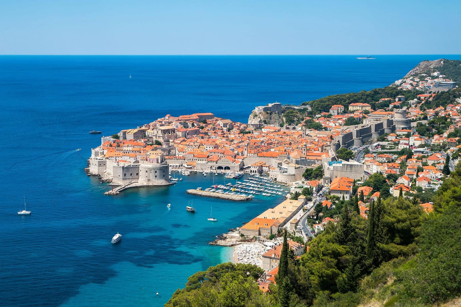 Elevated view of UNESCO-listed Dubrovnik Old Town, Croatia, with iconic terracotta rooftops enclosed by massive medieval city walls, the Lovrijenac Fortress on its rocky outcrop, the Old Port filled with boats, and the Adriatic Sea glowing turquoise to sapphire blue — book a fee-free Croatia vacation rental on Houfy | Houfy — No Fees, Book Direct