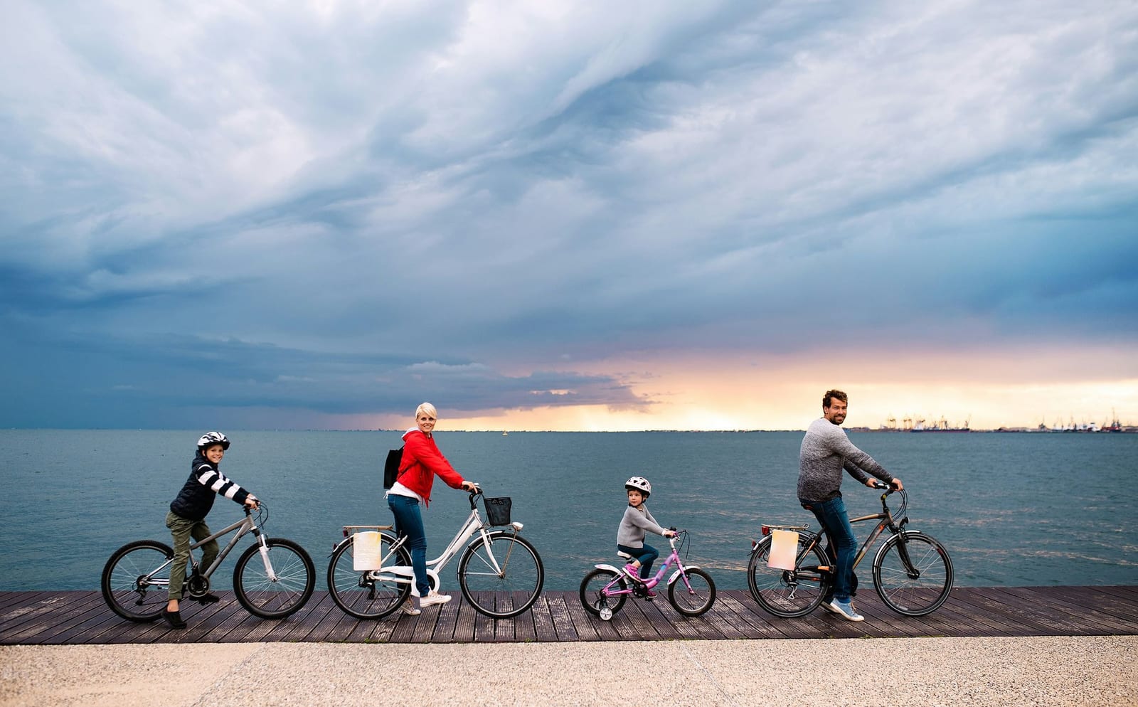 Family of four cycling along a coastal boardwalk at sunset under a dramatic stormy sky — book a fee-free cycling vacation rental on Houfy | Houfy — No Fees, Book Direct