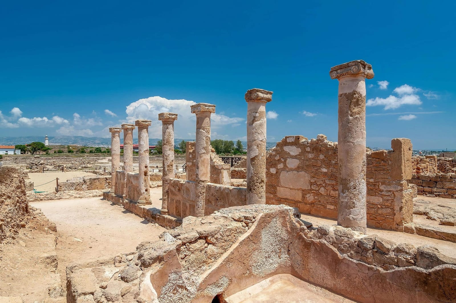 Seven ancient limestone columns of the House of Theseus at UNESCO-listed Paphos Archaeological Park, Cyprus, rising from sandy ochre earth and crumbling Roman-era stone foundations under a deep cerulean sky with white puffy clouds, with the white Paphos Lighthouse visible on the Mediterranean coastline in the distance — book a fee-free Cyprus vacation rental on Houfy | Houfy — No Fees, Book Direct