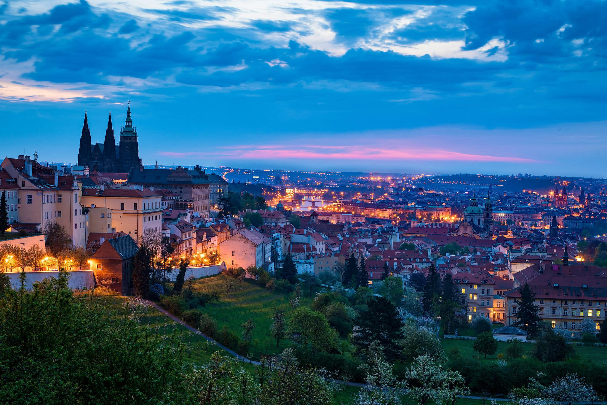 Blue hour panoramic view of Prague, Czechia, with the Gothic spires of St. Vitus Cathedral and Prague Castle silhouetted against a deep indigo sky, warm golden city lights glowing across a dense tapestry of red-tiled rooftops, and a lush green hillside with flowering white trees in the foreground — book a fee-free Czechia vacation rental on Houfy | Houfy — No Fees, Book Direct