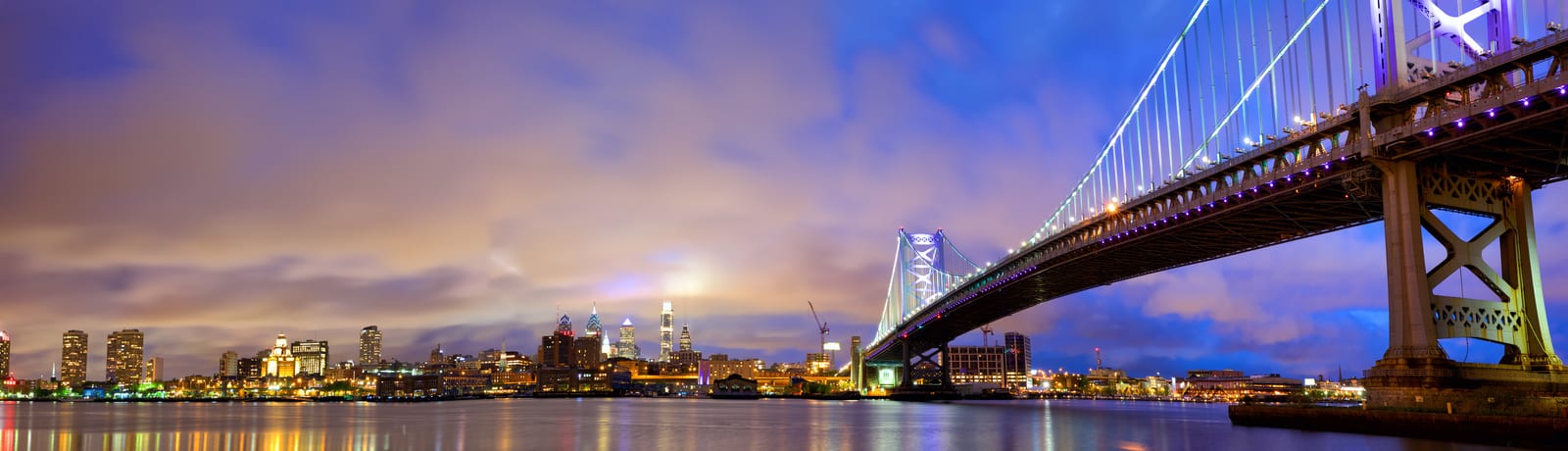 Philadelphia's iconic Ben Franklin Bridge illuminates the night sky, towering over the serene Delaware River. The vibrant city skyline with glowing skyscrapers accentuates the urban charm. | Houfy — No Fees, Book Direct