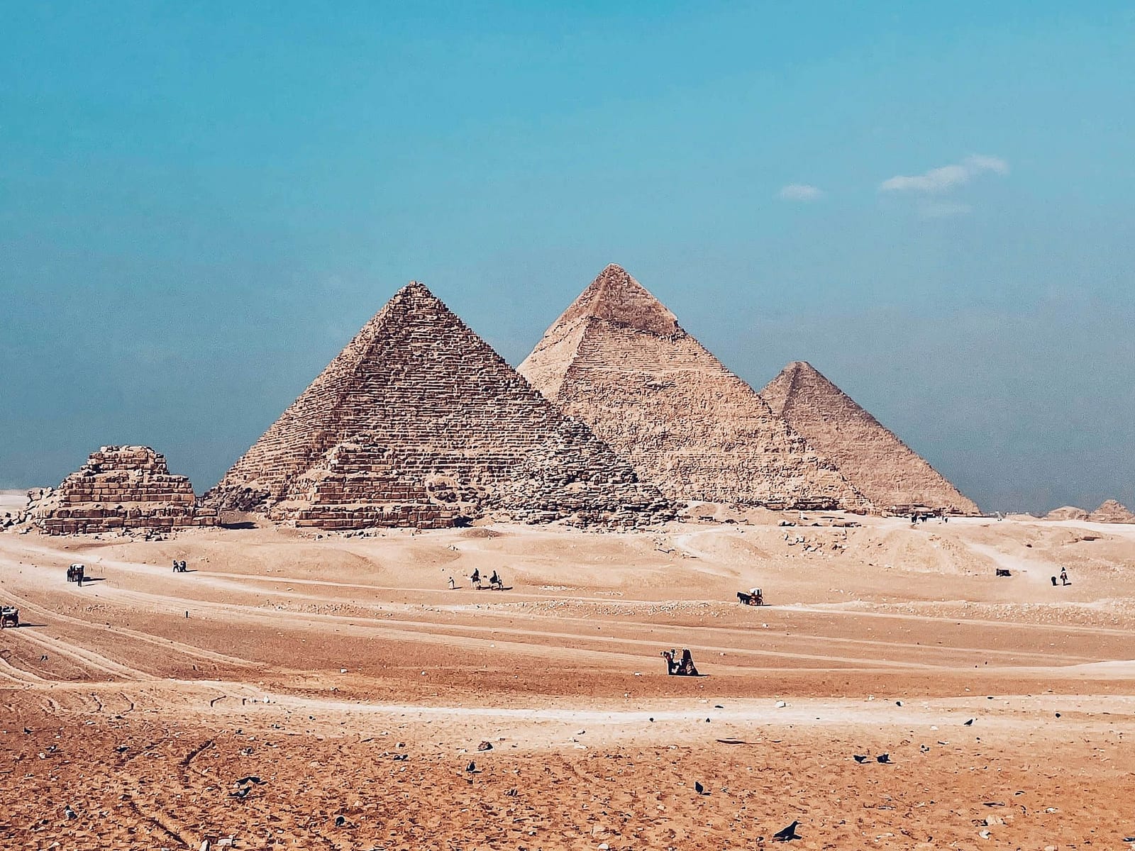 Midday panorama of the three Great Pyramids of Giza on the Cairo plateau, Egypt — the massive limestone-block pyramids of Khufu, Khafre, and Menkaure rising from undulating beige desert sand with small figures of tourists on camels and horses providing dramatic scale, against a saturated turquoise-blue sky — book a fee-free Egypt vacation rental on Houfy | Houfy — No Fees, Book Direct