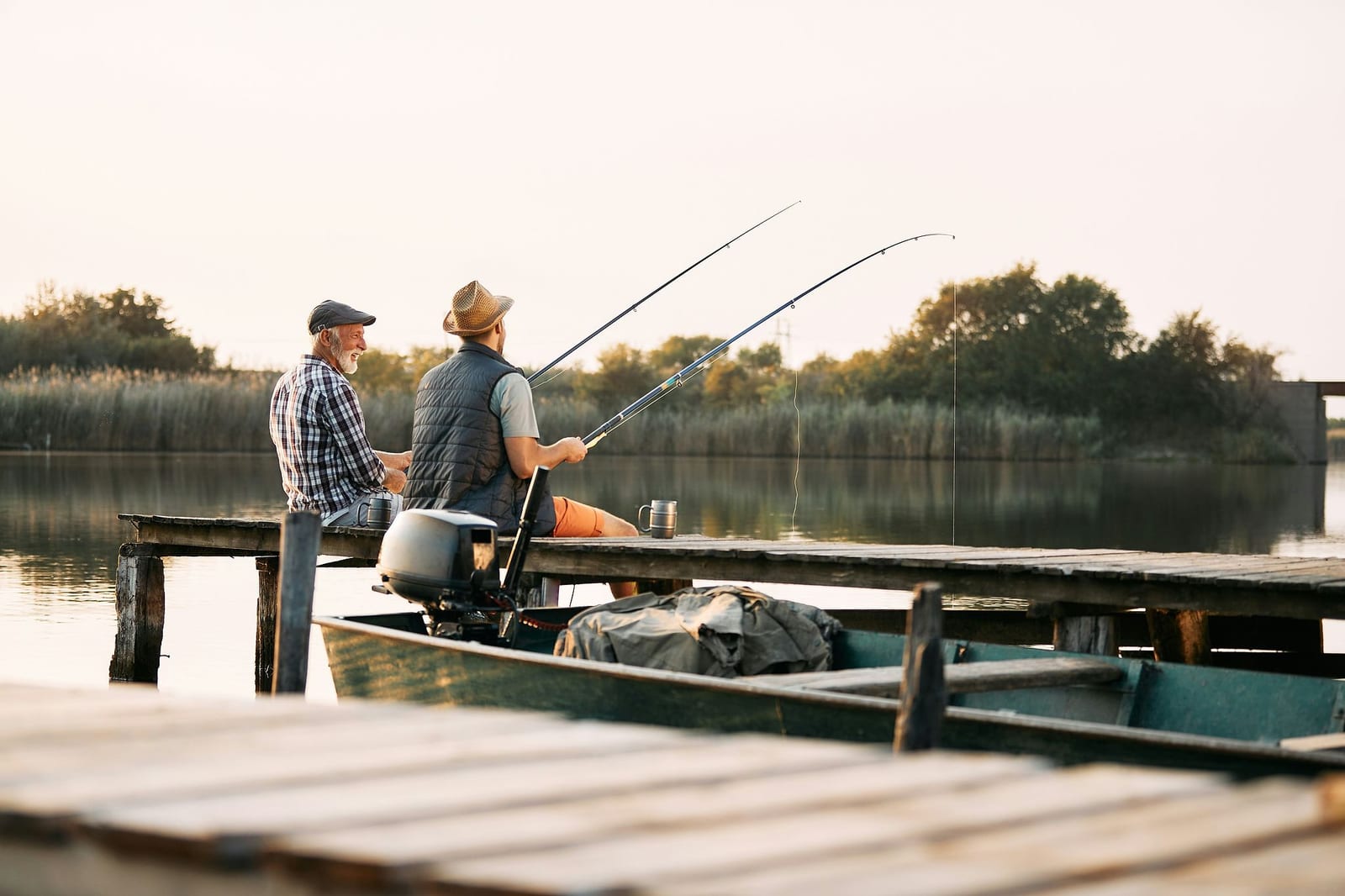 Two generations of anglers fishing side by side from a weathered wooden dock at golden hour, with a green fishing boat moored alongside and steaming mugs on the pier overlooking a glassy freshwater lake — book a fee-free fishing vacation rental on Houfy | Houfy — No Fees, Book Direct