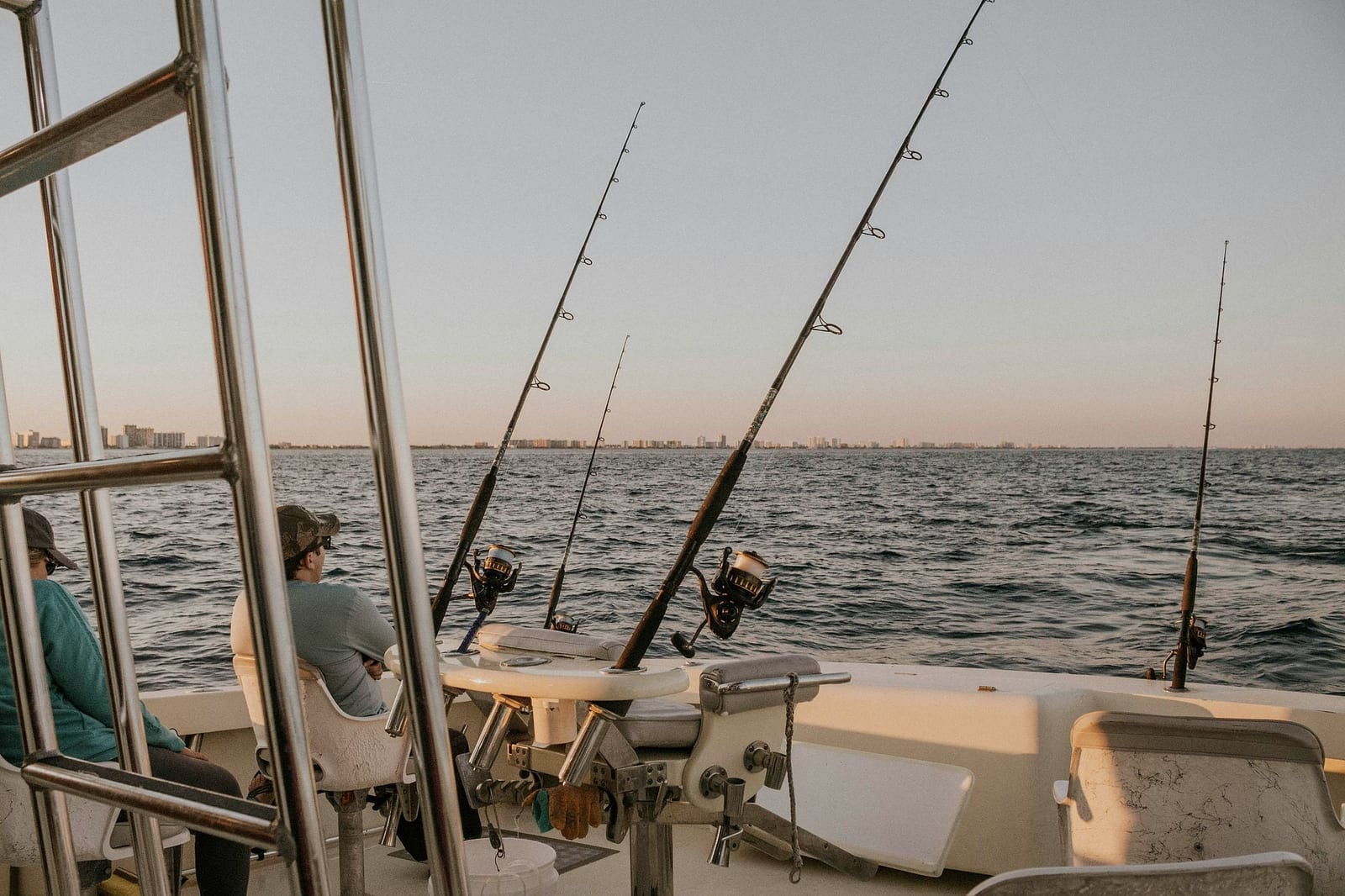 Two anglers relaxing on the stern deck of a deep-sea fishing charter boat at golden hour, with heavy-duty rods and reels set in rod holders and a coastal city skyline glowing on the horizon — book a fee-free fishing charter vacation rental on Houfy | Houfy — No Fees, Book Direct