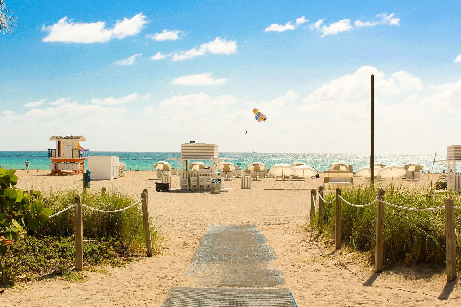Sunny South Beach, Miami with a classic Art Deco lifeguard stand, white beach umbrellas, sea oat dunes, and turquoise Atlantic Ocean waters under a brilliant blue Florida sky — book a fee-free Florida vacation rental on Houfy | Houfy — No Fees, Book Direct
