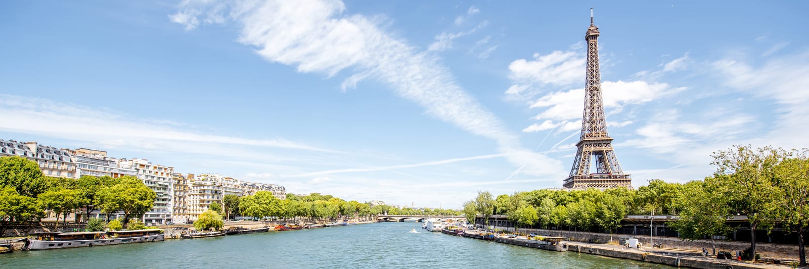 The Eiffel Tower rises majestically by the Seine River in Paris, framed by lush trees and vibrant houseboats. Clear blue sky and distinctive iron latticework capture the essence of this iconic cityscape. | Houfy — No Fees, Book Direct