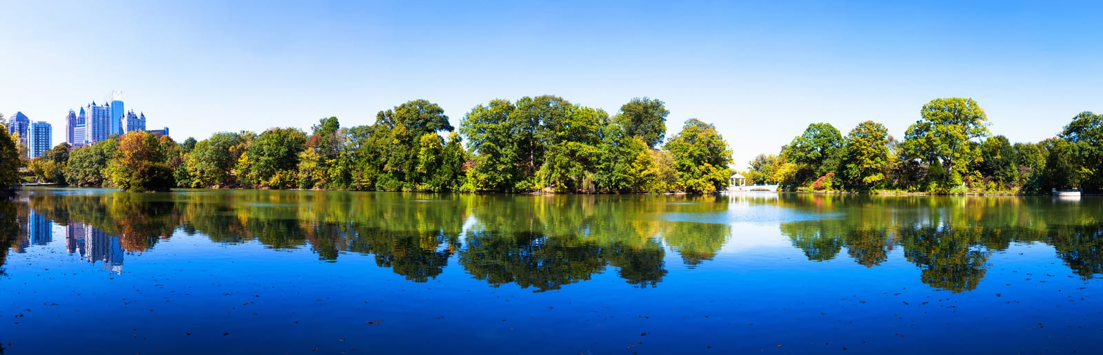Downtown Atlanta skyline rises beyond lush trees reflected in the calm waters of Piedmont Park's lake, capturing a serene urban oasis with clear blue skies above. | Houfy — No Fees, Book Direct