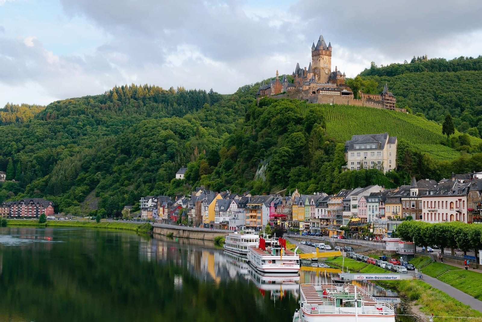 Panoramic view of Reichsburg Cochem castle rising above terraced Moselle Valley vineyards and the colorful half-timbered riverfront town of Cochem, Germany, with white river cruise ships docked along the calm Moselle River — book a fee-free Germany vacation rental on Houfy | Houfy — No Fees, Book Direct