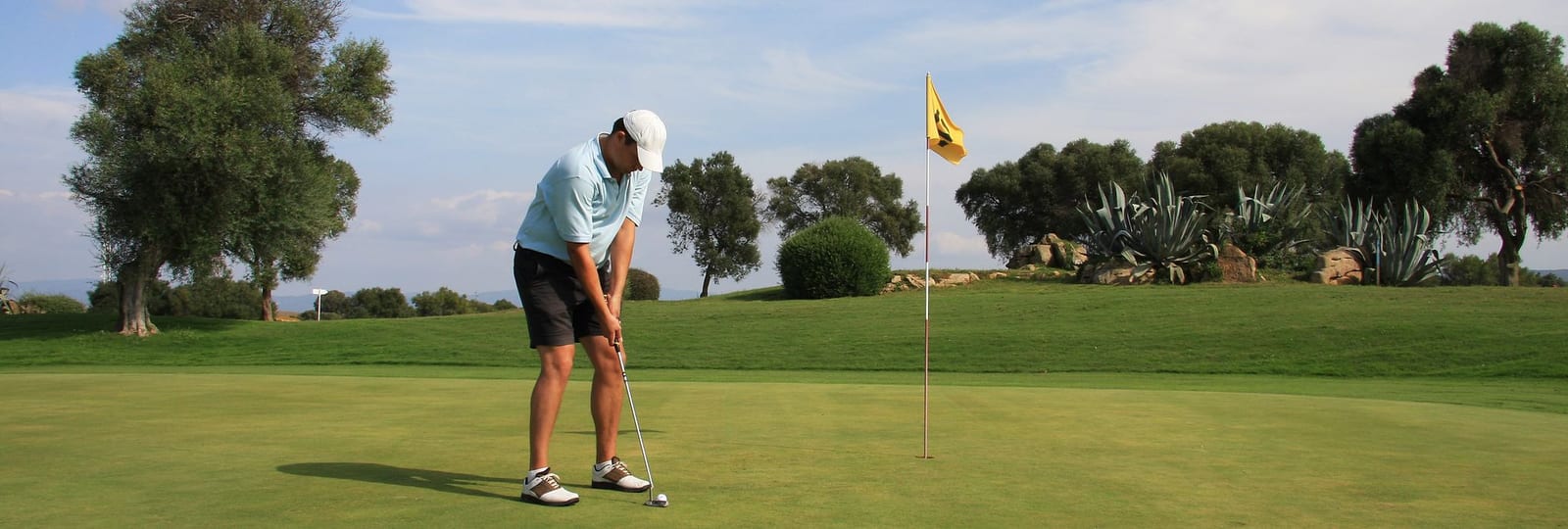 Golfer in focused putting stance on a pristine manicured green at a luxury resort course, with a yellow flag, agave succulents, olive trees, and soft blue mountain ranges under a clear afternoon sky — book a fee-free golf vacation rental on Houfy | Houfy — No Fees, Book Direct