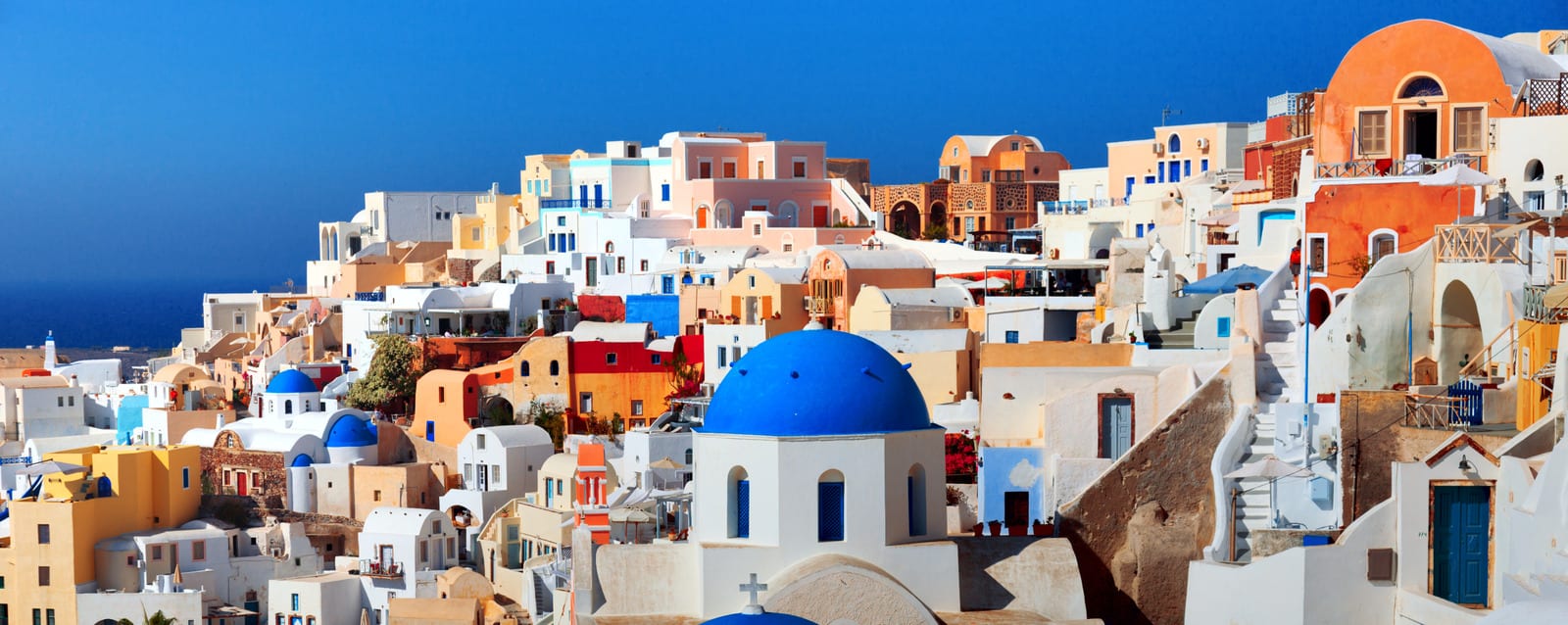 Panoramic view of Oia village on Santorini, Greece, with iconic blue-domed Orthodox churches, whitewashed cave houses in terracotta, ochre, and dusty rose, winding staircases, and the deep sapphire Aegean Sea under a cloudless azure sky — book a fee-free Greece vacation rental on Houfy | Houfy — No Fees, Book Direct