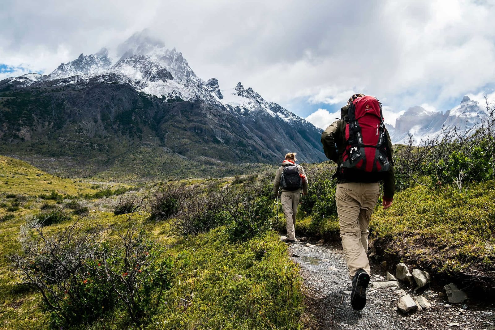 Two backpackers with heavy multi-day packs trekking a rugged narrow trail through a lush sub-alpine valley toward massive snow-capped granite peaks shrouded in clouds — book a fee-free hiking vacation rental on Houfy | Houfy — No Fees, Book Direct
