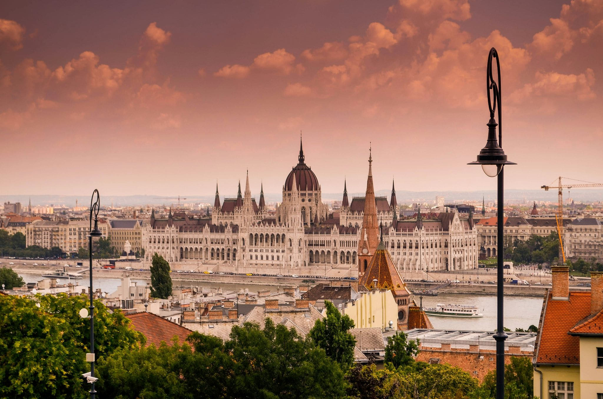 Golden hour panorama of Budapest, Hungary, with the Neo-Gothic Hungarian Parliament Building glowing in warm amber light, its grand central dome and symmetrical spires reflected in the wide Danube River, terracotta rooftops cascading down the Buda hillside, lush green trees framing the foreground, and a dramatic sky of billowing pink and dusty rose clouds at sunset — book a fee-free Hungary vacation rental on Houfy | Houfy — No Fees, Book Direct