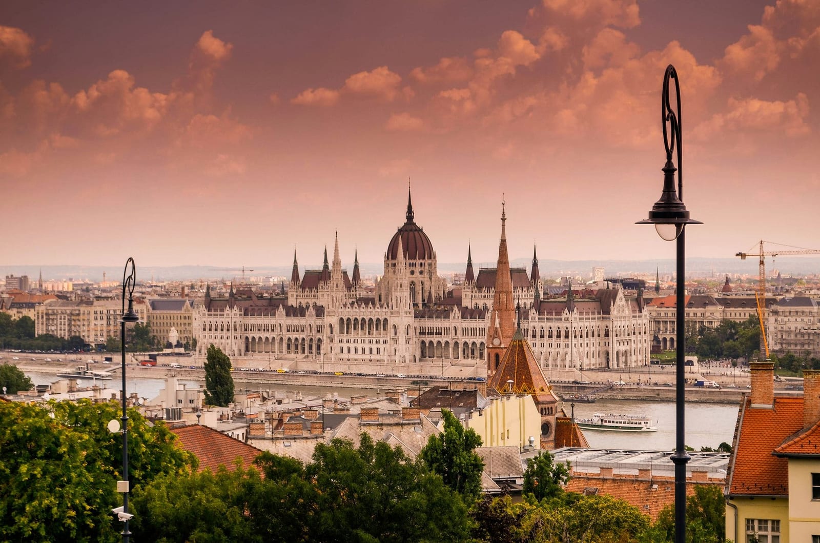 Golden hour panorama of Budapest, Hungary, with the Neo-Gothic Hungarian Parliament Building glowing in warm amber light, its grand central dome and symmetrical spires reflected in the wide Danube River, terracotta rooftops cascading down the Buda hillside, lush green trees framing the foreground, and a dramatic sky of billowing pink and dusty rose clouds at sunset — book a fee-free Hungary vacation rental on Houfy | Houfy — No Fees, Book Direct