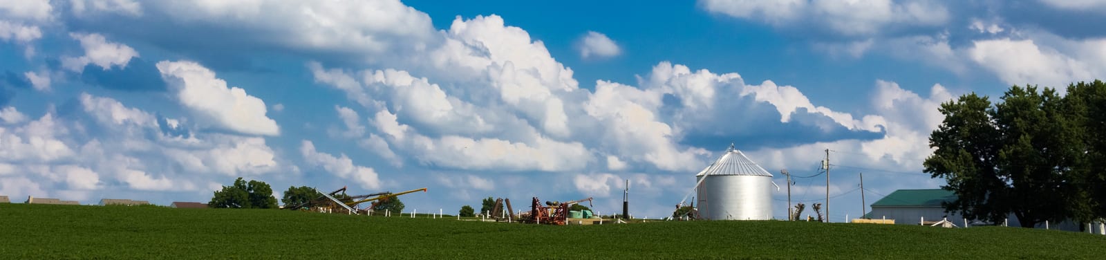 Expansive fields stretch under a vast sky with fluffy clouds, complemented by a large metal silo and distant farm machinery. This quintessential Midwestern landscape encapsulates rural charm. | Houfy — No Fees, Book Direct
