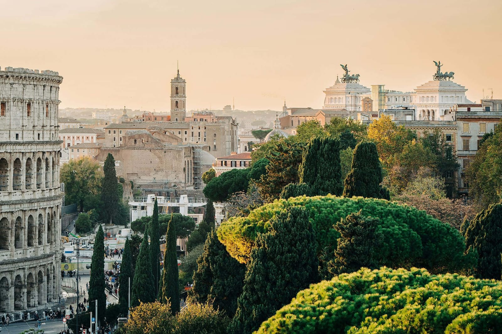 Golden hour panorama of Rome, Italy, with the iconic Colosseum, Roman Forum ruins, and the white marble Altare della Patria framed by lush Italian stone pines and cypress trees under a warm peach and amber sky — book a fee-free Italy vacation rental on Houfy | Houfy — No Fees, Book Direct