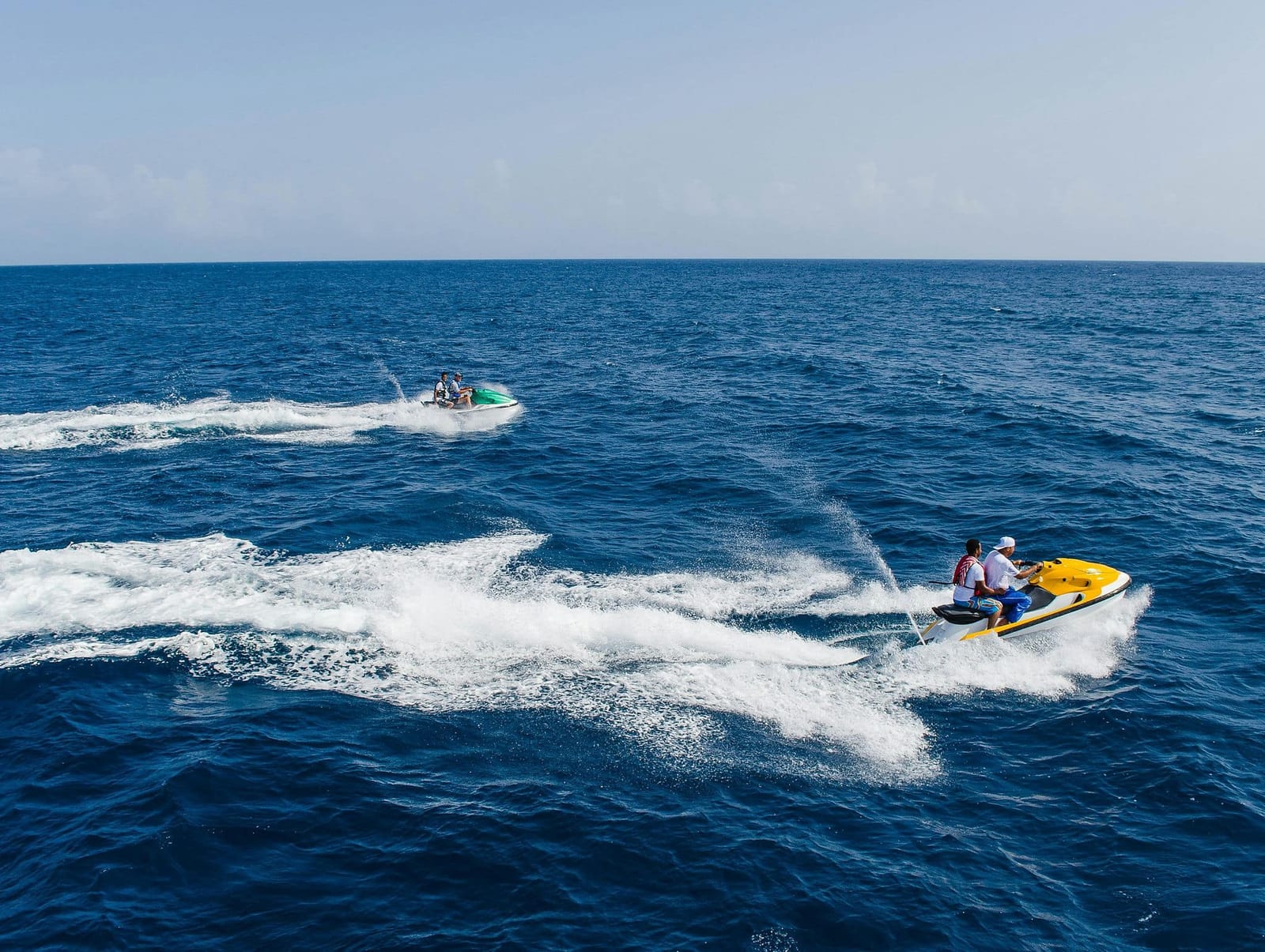 Two riders on a yellow jet ski cutting through deep blue open ocean at high speed, trailing a dramatic white wake, with a second green jet ski following in the distance — book a fee-free jet skiing vacation rental on Houfy | Houfy — No Fees, Book Direct