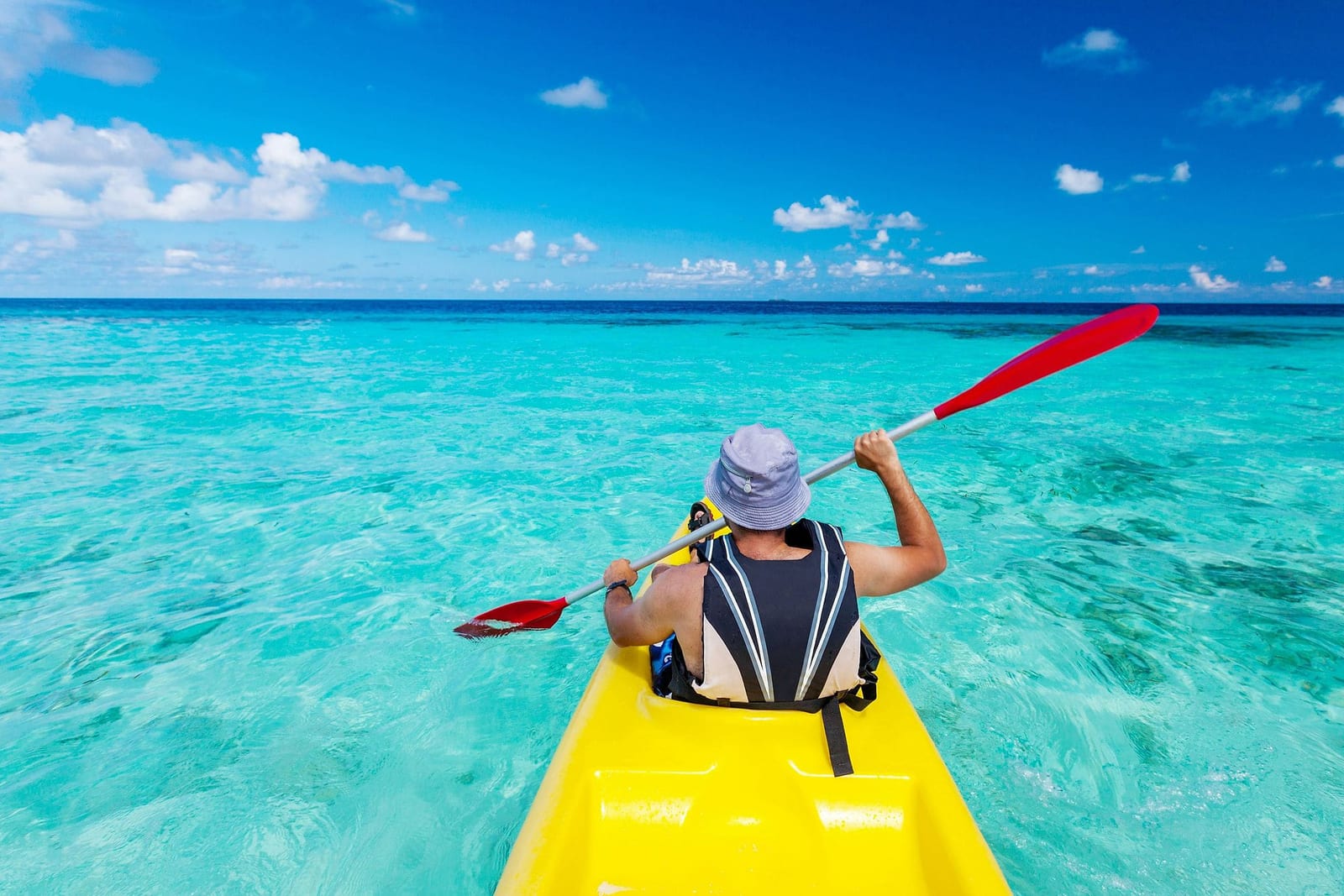 POV shot of a kayaker in a bright yellow sit-on-top kayak with a red-bladed paddle gliding across crystal-clear turquoise tropical ocean water with coral and sandy seabed visible below under a vivid blue sky — book a fee-free kayaking vacation rental on Houfy | Houfy — No Fees, Book Direct