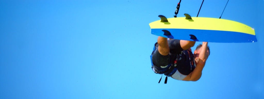 Kitesurfer launching a high big-air jump against a cloudless vivid blue sky, with a bold yellow and royal blue twin-tip kiteboard and water droplets catching the light at peak height — book a fee-free kitesurfing vacation rental on Houfy | Houfy — No Fees, Book Direct