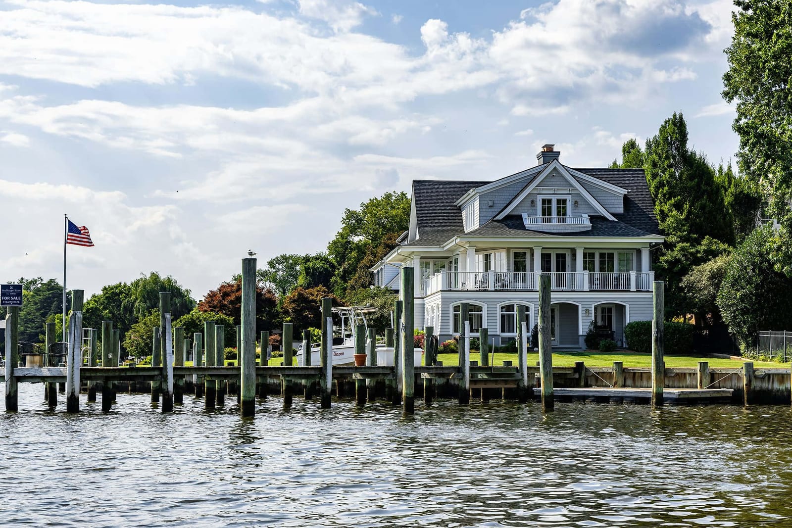 Grand three-story lakefront vacation home with grey cedar shingle siding, white-trimmed multi-level balconies, and arched ground-floor patio, set on a manicured green lawn above a weathered wooden dock with tall vertical pilings extending into dark rippling lake water, an American flag on a tall flagpole, and a white powerboat moored at the shore under a vivid blue sky with voluminous white cumulus clouds — book a fee-free lakefront vacation rental on Houfy | Houfy — No Fees, Book Direct